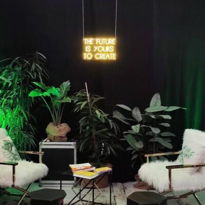 Zwei weiße Stühle aus Kunstfell stehen sich in einem Raum mit Topfpflanzen, schwarzen Wänden und grüner Beleuchtung gegenüber. Auf einer Leuchtreklame darüber steht THE FUTURE IS YOURS TO CREATE. Auf einem Tisch zwischen den Stühlen liegen Bücher.