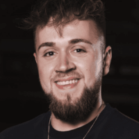 Ein junger Mann mit kurzen, gewellten braunen Haaren und einem Bart lächelt in die Kamera. Er trägt ein dunkles Hemd und eine Halskette. Hinter ihm ist ein dunkler, unscharfer Hintergrund zu sehen.