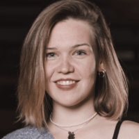 Eine Frau mit schulterlangem hellbraunem Haar, die ein graues schulterfreies Oberteil, Reifohrringe, eine Perlenkette und einen schwarzen Anhänger trägt, lächelt vor einem dunklen Hintergrund in die Kamera.