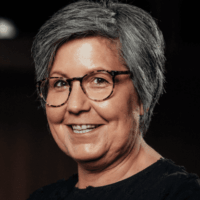 Eine Person mittleren Alters mit kurzen grauen Haaren, die eine Brille und ein dunkles Oberteil trägt, lächelt in die Kamera vor einem dunklen Hintergrund.