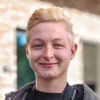 Eine Person mit kurzem, blondem Haar lächelt in die Kamera. Sie trägt ein dunkles Oberteil und steht im Freien vor einem unscharfen Hintergrund mit einem Gebäude und Fenstern.