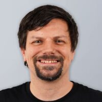 Ein Mann mit kurzen dunklen Haaren, einem Schnurrbart und einem Spitzbart lächelt. Er trägt ein schwarzes Hemd und steht vor einem schlichten hellgrauen Hintergrund.