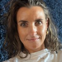 Eine Frau mit langen braunen Haaren und hellen Augen trägt ein weißes Oberteil und goldene Ohrringe und blickt direkt in die Kamera. Der Hintergrund ist dunkelblau und hat ein strukturiertes Aussehen.