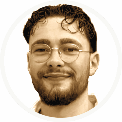 Ein Mann mit kurzen dunklen Haaren, einer Brille und einem Bart lächelt leicht. Er ist von den Schultern aufwärts vor einem schlichten weißen Hintergrund abgebildet.