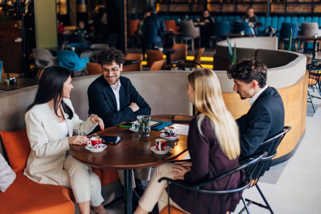 Vier Personen in Geschäftskleidung sitzen um einen runden Tisch in einem modernen Café und unterhalten sich. Auf dem Tisch stehen Kaffeetassen, Gläser und ein Telefon. Andere Gäste sind im Hintergrund zu sehen.