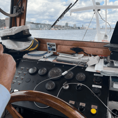 Blick aus dem Steuerhaus eines Schiffes mit Steuerrad, Navigationsinstrumenten, einer Kapitänsmütze auf dem Armaturenbrett, verschiedenen Papieren und Geräten sowie einer Hand, die das Steuerrad ergreift, wobei das Wasser und die Skyline der Stadt durch das Fenster zu sehen sind.